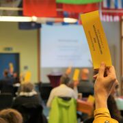 gehobene Hand mit Stimmzettel im Vordergrund; dahinter verschwommen mehrere Personen im Raum, die auch Stimmzettel nach oben halten